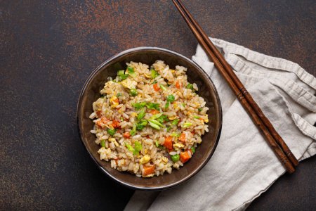 Authentic Chinese and Asian fried rice with egg and vegetables in ceramic brown bowl top view on dark rustic concrete table background. Traditional dish of China