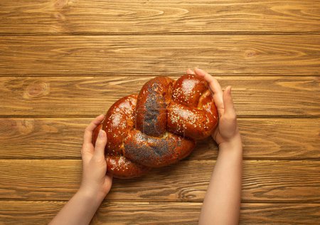 Photo for Female hands holding freshly baked Challah bread covered with poppy and sesame seeds, top view on rustic wooden background, traditional festive Jewish cuisine. - Royalty Free Image
