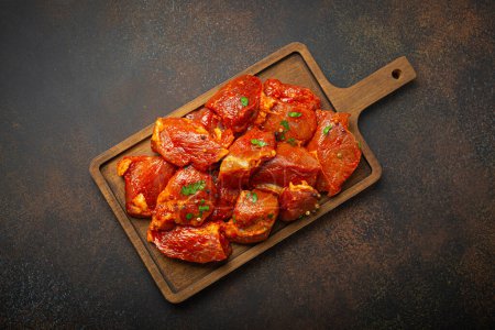 Photo for Raw uncooked chopped pieces of pork marinated with seasonings and parsley on wooden cutting board top view on dark rustic background. Cooking meal with marinated pork fillet pieces. - Royalty Free Image