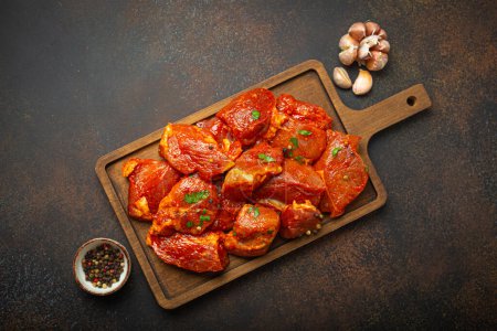 Photo for Raw uncooked chopped pieces of pork marinated with seasonings and parsley on wooden cutting board top view on dark rustic background. Cooking meal with marinated pork fillet pieces. - Royalty Free Image