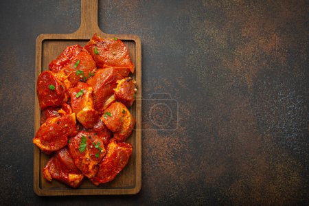 Photo for Raw uncooked chopped pieces of pork marinated with seasonings and parsley on wooden cutting board top view on dark rustic background. Cooking meal with marinated pork fillet pieces. Space for text. - Royalty Free Image
