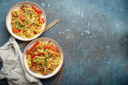 Zwei Schüsseln mit Chow Mein oder Lo Mein, traditionelle chinesische Rührnudeln mit Fleisch und Gemüse, serviert mit Essstäbchen von oben auf rustikalem blauem Betonhintergrund, Platz für Text.