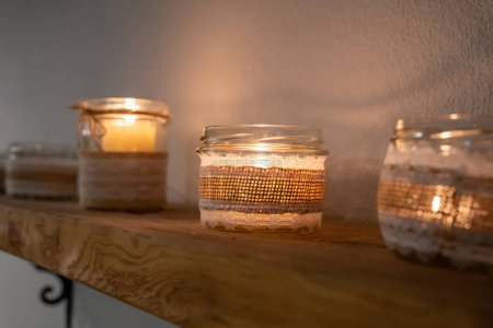 Photo for Lit candles in glass jars with fabric on wooden shelf - Royalty Free Image