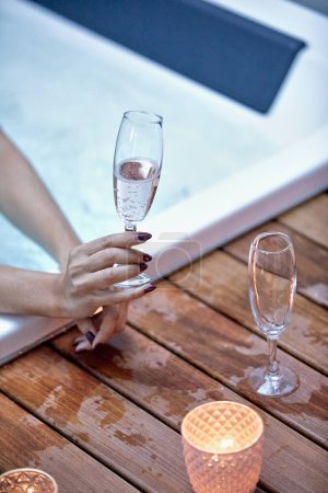 Elegante Hand hält eine Champagnerflöte an einem Whirlpool mit einem leeren Glas und Votivkerzen auf einem Holzdeck.