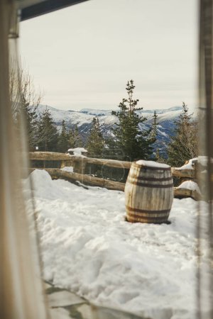 Snowy mountain view through a cabin window, with a wooden barrel.