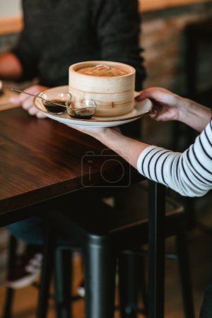 Téléchargez les photos : Serveuse servant un homme à une table de restaurant avec une assiette comportant deux petits récipients à sauce et un grand bol en bois de gyozas - en image libre de droit