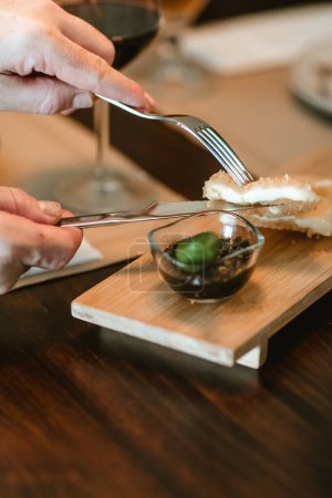 Close-up of a man's hands holding a knife and fork, cutting a piece of breaded food on a wooden table.