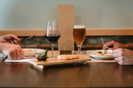 zwei Männerhände, ein Weinglas, ein Bier, ein Restaurantmenü und ein Holzteller mit paniertem Essen, jeder Mann hält Utensilien bereit, um zu essen