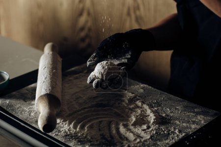 Close-up of a chef carefully dusting a dessert with flour, one hand holding the dessert, with a tray of flour and a rolling pin in the background.