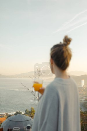 Sunrise Serenity: Mujer en suéter de gran tamaño disfrutando de un vaso de jugo con vistas a la costa Sunrise
