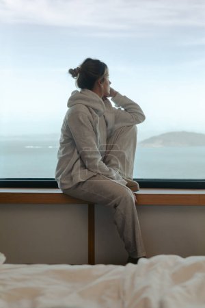 Mujer joven contemplativa mirando al mar desde la repisa de una ventana de una habitación moderna en ropa de salón cómoda
