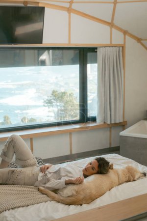 Coastal Comfort: Relaxed Woman Lying on Bed with Her Dog Enjoying the Ocean View from a Stylish Room