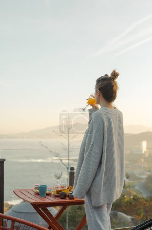 Seaside Morning Bliss : Femme sirotant du jus d'orange profitant du lever du soleil sur l'océan depuis une terrasse panoramique