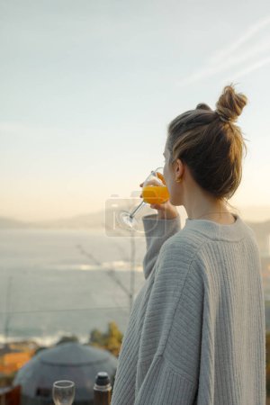 Elegancia de la mañana crujiente: Mujer joven disfrutando de un vaso de jugo de naranja fresco en una terraza con vistas al mar al amanecer