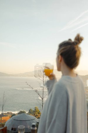 Sunrise Serenity: Frau im übergroßen Pullover genießt ein Glas Saft mit Blick auf den Sonnenaufgang an der Küste