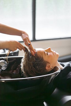 Eine Frau lehnt an einer Shampoo-Station im Salon, während ein Friseur ihre Kopfhaut mit einer Haarbehandlung massiert. Die Szene spielt in einem modernen Salon mit natürlichem Licht.