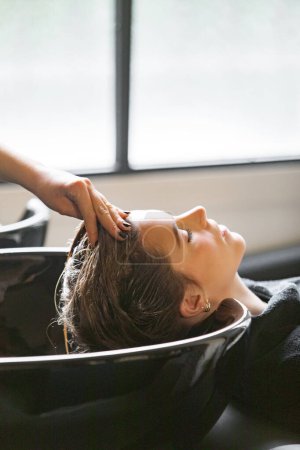 A woman enjoys a relaxing scalp massage at a salon shampoo station. The scene captures a modern salon setting with soft natural lighting.