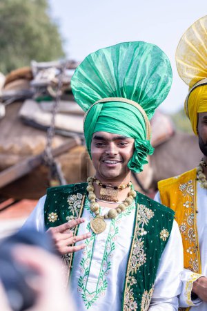 Foto de Bikaner, Rajasthan, India - January 2023: Punjabi Bhangra, Portrait of young sikh male in traditional punjabi colorful dress and turban performing bhangra dance with smile in camel festival with focus - Imagen libre de derechos