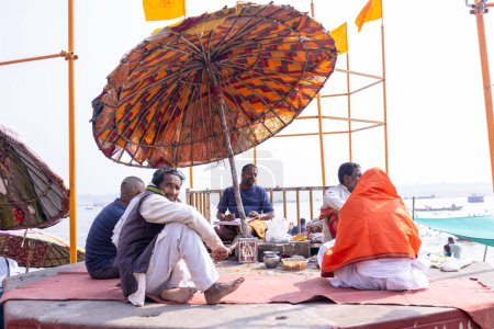 Foto de Varanasi, Uttar Pradesh, India - Noviembre de 2022: Retrato de un sacerdote brahmán indio no identificado sentado bajo un paraguas en ghat cerca de bandas fluviales en la ciudad de varanasi. Sacerdotes realizaron rituales en ghats. - Imagen libre de derechos