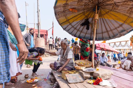 Foto de Varanasi, Uttar Pradesh, India - Noviembre de 2022: Retrato de un sacerdote brahmán indio no identificado sentado bajo un paraguas en ghat cerca de bandas fluviales en la ciudad de varanasi. Sacerdotes realizaron rituales en ghats. - Imagen libre de derechos