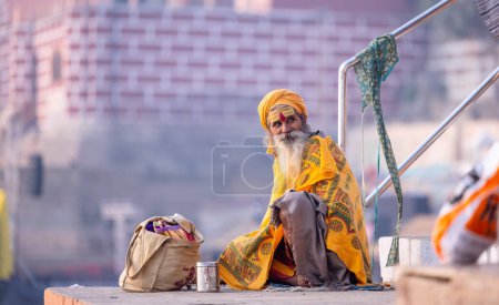 Téléchargez les photos : Varanasi, Uttar Pradesh, Inde - Novembre 2022 : Portrait du vieux sadhu indien non identifié baba assis sur des escaliers colorés sur des ghats près de la rivière Ganges dans la ville de varanasi en tenue traditionnelle. - en image libre de droit
