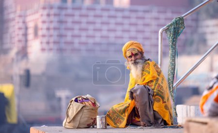 Foto de Varanasi, Uttar Pradesh, India - Noviembre 2022: Retrato de un viejo sadhu baba indio no identificado sentado en coloridas escaleras en ghats cerca de las bandas fluviales en la ciudad de varanasi con vestimenta tradicional. - Imagen libre de derechos