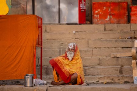 Foto de Varanasi, Uttar Pradesh, India - 20 de noviembre de 2022: Retrato de un hombre sadhu sagrado indio no identificado sentado cerca de las pandillas del río en la ciudad de varanasi con vestimenta tradicional. - Imagen libre de derechos