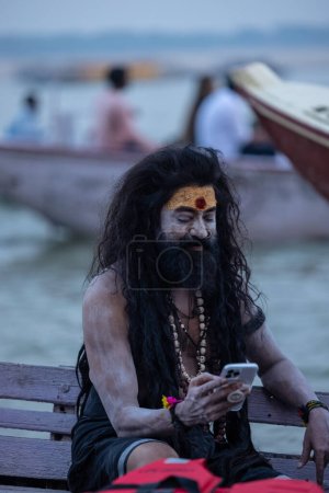 Foto de Varanasi, Uttar Pradesh, India - 20 de noviembre de 2022: Retrato de un actor indio no identificado como naga sadhu con preguntar en su rostro y cuerpo sentado en un barco de madera en las bandas fluviales en varanasi. - Imagen libre de derechos