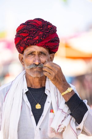 Téléchargez les photos : Pushkar, Rajasthan, Inde - 05 novembre 2022 : Foire de Pushkar, portrait d'un vieil homme rajasthani vêtu d'une robe traditionnelle blanche et d'un turban coloré touchant sa moustache à la foire de pushkar. - en image libre de droit