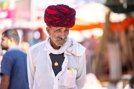 Foto de Pushkar, Rajastán, India - 05 de noviembre 2022: Feria de Pushkar, Retrato de un hombre viejo rajasthani con vestido tradicional blanco y turbante colorido tocándose el bigote en el recinto ferial de pushkar. - Imagen libre de derechos