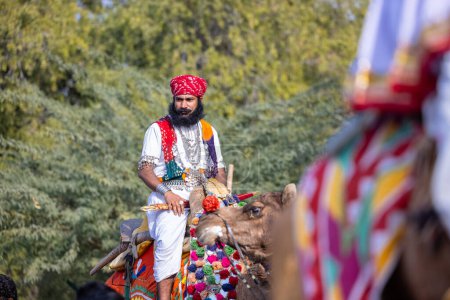 Bikaner, Rajasthan, India - January 13 2023: Camel Festival, Portrait of an young rajasthani male with beard and moustache wearing white traditional rajasthani dress and turban riding on camel.