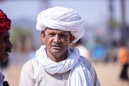 Téléchargez les photos : Pushkar, Rajasthan, Inde - 06 Novembre 2022 : Foire de Pushkar, Portrait d'un vieil homme rajasthani avec robe traditionnelle blanche et turban coloré jouissant au terrain de foire de pushkar - en image libre de droit