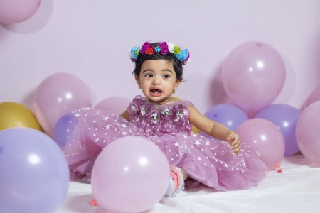 Photo for First year's birthday. Happy little girl in pink dress and flowery hair band sits in background with garlands and pink balloons, celebrating her first birthday. Decoration birthday. copy space. - Royalty Free Image