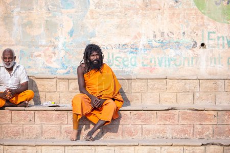 Foto de Varanasi, Uttar Pradesh, India - 05 de marzo de 2023: Retrato del viejo sadhu baba sagrado con ropa tradicional sentado en los ghats cerca de las pandillas en varanasi. Kashi es la ciudad más antigua y santa. - Imagen libre de derechos