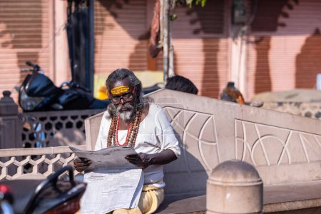 Photo for Varanasi, Uttar Pradesh, India - March 05 2023: Portrait of old holy south indian brahmin man in traditional dress sitting on the ghats near ganges in varanasi during sunrise and reading newspaper - Royalty Free Image