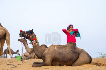Foto de Retrato de una joven india rajasthani con colorido vestido tradicional llevando camello en Pushkar Camel Fair ground durante la mañana brumosa de invierno en Pushkar, Rajasthan. - Imagen libre de derechos