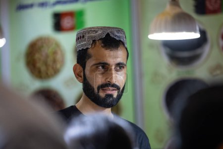 Foto de Nueva Delhi, India - 18 de noviembre de 2023: Retrato de un vendedor de ropa tradicional de Afganistán en un mostrador en la feria de Nueva Delhi para vender sus productos. - Imagen libre de derechos