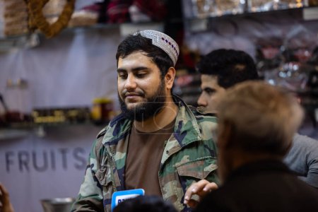 Foto de Nueva Delhi, India - 18 de noviembre de 2023: Retrato de un vendedor de ropa tradicional de Afganistán en un mostrador en la feria de Nueva Delhi para vender sus productos. - Imagen libre de derechos