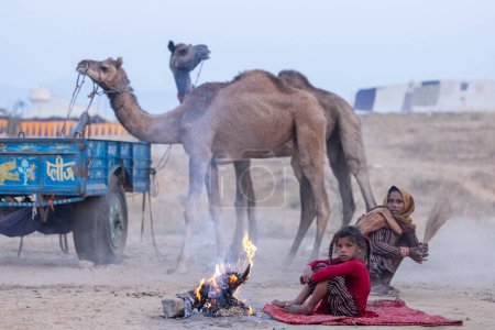 Foto de Pushkar, Rajastán, India - 24 de noviembre de 2023: Retrato de una joven refugiada abandonada sentada en el suelo cerca de una hoguera en la nebulosa mañana de invierno durante la feria de camellos de pushkar. - Imagen libre de derechos