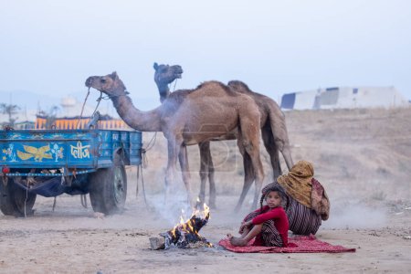 Photo for Pushkar, Rajasthan, India - November 24, 2023: Portrait of young abandon refugee girl sitting on ground near bonfire at foggy winter morning during pushkar camel fair. - Royalty Free Image