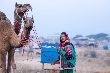 Foto de Pushkar, Rajasthan, India - November 24 2023: Portrait of an young Indian rajasthani woman in colorful traditional dress carrying camel at Pushkar Camel Fair ground during winter morning. - Imagen libre de derechos