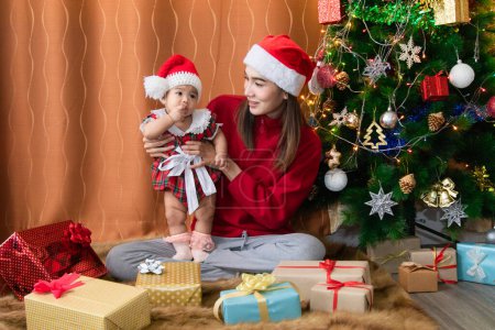 Asian happy young mother and adorable baby on sweater clothes sit near Christmas tree decoration play together at home in Holiday greeting season, beautiful mom with Santa red hat kissing sweet girl