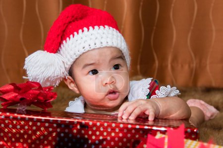 Asian adorable baby girl with Christmas red hat curiosity looking to present box, curiosity girl 6 month old try to remove wrapping gift with tiny finger. infant baby use fingers unwrap gift box