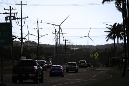 Foto de Kahuku, HI - 3 de enero de 2024: Turbinas eólicas renovables que generan energía eléctrica limpia en una noche nublada a las líneas eléctricas del vecindario hawaiano, mientras que los vehículos a gas pasan por la carretera - Imagen libre de derechos