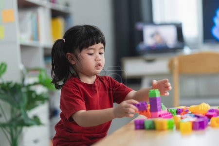 Asian cute funny preschooler little girl in a colorful shirt playing with lego or construction toy blocks building a tower in kindergarten room or living room. Kids playing. Children at day care.