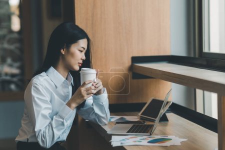 Photo for Asian freelance woman smiling holding cup of hot coffee and working on laptop computer on wooden table at cafe. Entrepreneur woman working for her business at coffee shop. Business work from anywhere - Royalty Free Image