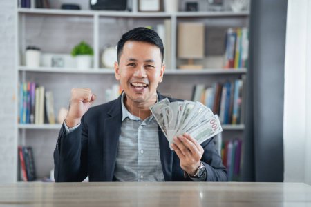 Téléchargez les photos : Costume homme d'affaires asiatique tenant billet de dollar et souriant heureux obtenir de l'argent assis bureau dans le salon au bureau à la maison. Épargner la richesse d'investissement concept. Succès pari casino en ligne ou investir pour le stock. - en image libre de droit