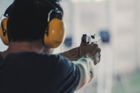 Photo for Asian shooter man wearing noise canceling headphones and black clothes practicing shooting short gun at the shooting range. Shooting sports for meditation and self-defense, recreational activities. - Royalty Free Image