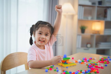 Fröhliches asiatisches Mädchen glücklich und lächelnd hebt die Hand beim Spielen bunter Lego-Spielzeuge, sitzt auf dem Tisch im Wohnzimmer, spielt kreativ mit Lego, baut bunte Strukturen Kreativität vorstellen.