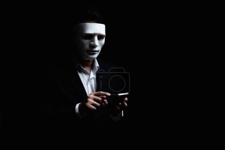 Unknown businessman wearing mask with covered face using mobile phone makes an anonymous call intimidating and threatening the interlocutor on dark background. hacker callcenter concept.